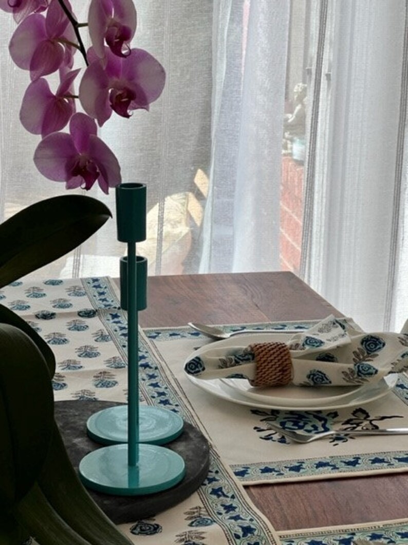May include: A teal blue metal candle holder with a round base sits on a table setting with a white tablecloth and blue floral pattern. A woven napkin ring is on a white plate.