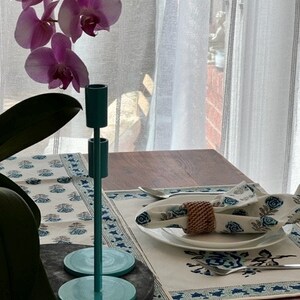 May include: A teal blue metal candle holder with a round base sits on a table setting with a white tablecloth and blue floral pattern. A woven napkin ring is on a white plate.