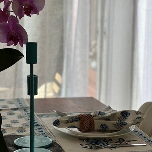 May include: A teal metal candle holder with two arms, sitting on a table with a white tablecloth and a place setting. The tablecloth has a blue and white floral pattern.