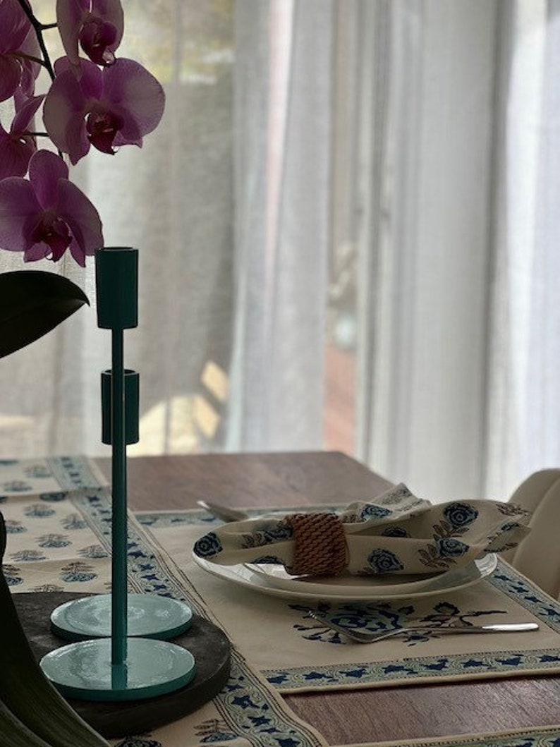 May include: A teal metal candle holder with two arms, sitting on a table with a white tablecloth and a place setting. The tablecloth has a blue and white floral pattern.