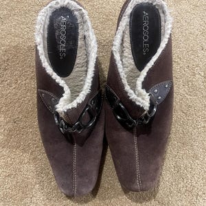 May include: A pair of brown suede Aerosoles slip-on shoes with white faux fur lining. The shoes have a decorative black chain and rivet detail. The brand name "Aerosoles" is visible on the insole.