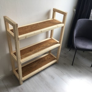 以下が含まれることがあります： 自然な木の棚と明るい木製のフレームを持つ3段の木製棚ユニット。棚は濃い茶色に着色されています。棚ユニットは明るい木製の床の上に立っています。