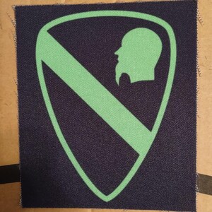 Puede incluir: Parche azul marino con un diseño de escudo verde. Dentro del escudo hay una franja diagonal verde y una silueta verde del perfil de un hombre barbudo. El parche tiene una apariencia texturizada.