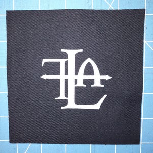 Peut inclure: Un écusson carré en tissu bleu marine foncé avec un monogramme blanc des lettres "T", "L", "A" et "E". Les lettres sont entrelacées avec des flèches horizontales.