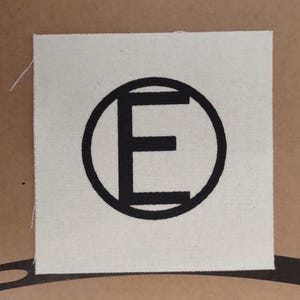 May include: Square, off-white fabric patch with a black circular border and a bold, black letter "E" in the center. The fabric has a slightly textured appearance. The patch is likely for sewing or ironing onto clothing or accessories.
