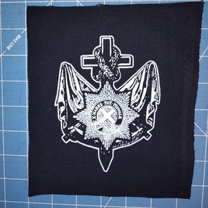May include: Black square patch with a white emblem featuring a cross, knot, and starburst design. The patch has a raw edge and is likely for sewing or ironing onto clothing or accessories.
