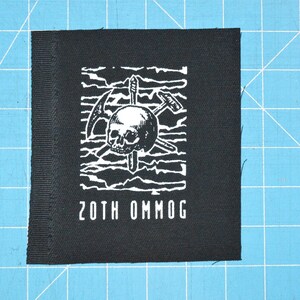 Pode incluir: Remendo preto com um design de caveira branca e picaretas cruzadas. O texto "20TH OMMOG" é impresso abaixo do design.