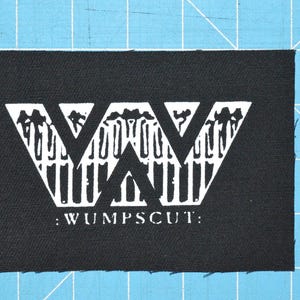 Pode incluir: Remendo preto com um gráfico branco de um "W" estilizado com o texto ":WUMPSCUT:" abaixo.