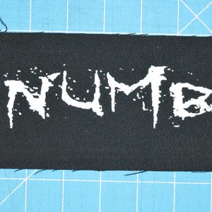Pode incluir: Patch de tecido preto com letras brancas que dizem "NUMB".