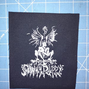 Pode incluir: Patch quadrado preto com um gráfico branco de uma figura alada e o nome da banda "SKINNY PUPPY" em uma fonte estilizada. Adequado para roupas ou acessórios.