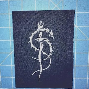 Pode incluir: Um patch de tecido azul escuro com uma ilustração branca estilizada de uma criatura semelhante a um dragão coroado. O desenho está centrado no patch, que tem uma borda ligeiramente desfiada. O fundo é uma grelha azul.