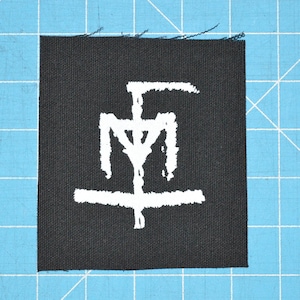 Pode incluir: Um patch de tecido preto quadrado com um símbolo branco estilizado. O símbolo é uma combinação de linhas verticais e horizontais, com uma forma de cruz na parte inferior. O patch está sobre uma superfície de grade azul.