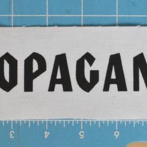 Può includere: Toppa di tessuto bianco con la parola "PROPAGANDA" stampata in lettere maiuscole nere.