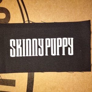 Pode incluir: Um patch retangular preto com o texto branco "SKINNY PUPPY". O patch tem bordas desgastadas e é colocado contra um fundo marrom com formas geométricas marrom escuro.