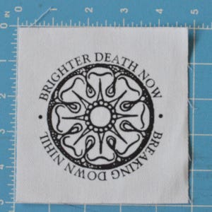 May include: A white fabric patch with black text and a black and white graphic design. The text reads "Breaking Down Nihil Brighter Death Now".