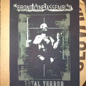 Może przedstawiać: Czarna naszywka z napisem "FRONT LINE ASSEMBLY" w kolorze białym. Poniżej biało-szara grafika postaci, z napisem "TOTAL TERROR" wydrukowanym poniżej w kolorze białym.