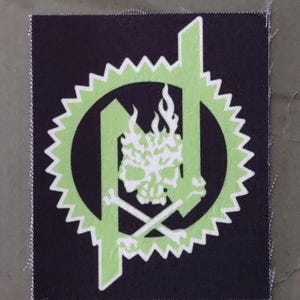 Puede incluir: Un parche cuadrado con fondo negro presenta un diseño verde neón. El diseño incluye una calavera con llamas, huesos cruzados y un borde en forma de engranaje. El parche tiene un borde en zigzag blanco.