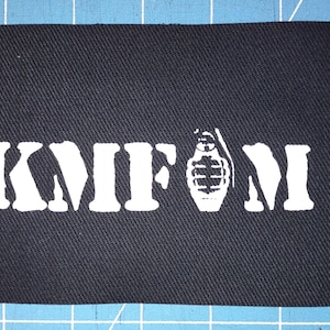 May include: A black rectangular patch with white block letters spelling "KMFDM" and a white graphic of a hand grenade. The patch is made of a textured fabric and is likely an accessory for clothing or bags.