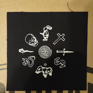 Op de afbeelding: Zwart vierkantige patch met witte geborduurde ontwerpen, waaronder een schedel, kruis, mes, dollarteken en een konijnachtig figuur. Het woord "swans" is ook geborduurd. Voor kleding of accessoires.