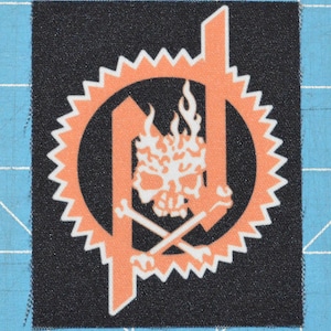Pode incluir: Um remendo bordado preto e laranja com um design de caveira e ossos cruzados. A caveira é cercada por chamas e o design é fechado em um círculo com uma borda dentada.