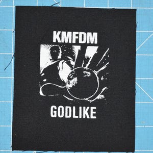 May include: Black and white embroidered patch with the text "KMFDM GODLIKE" and a graphic of a person holding a sphere.