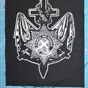 Pode incluir: Patch preto e branco com um design de âncora branca. A âncora tem uma cruz no topo e um círculo com o texto "X Marks The Peddler" no centro.