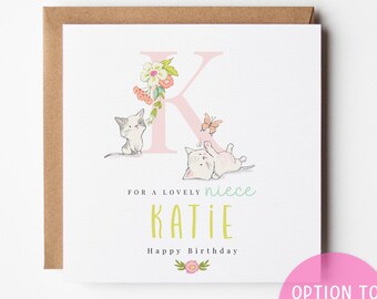 May include: A greeting card with a brown envelope. The card features a pink letter "K" with floral accents, two cat illustrations, and the text "For a lovely niece Katie Happy Birthday". A pink circle with the text "Option to include age" is in the bottom right.