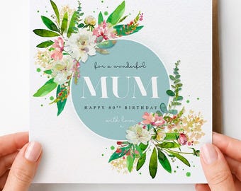 May include: A square greeting card featuring a central light blue circle with the text "For a wonderful MUM HAPPY 80TH BIRTHDAY with love". The card is decorated with watercolor illustrations of green leaves and pink and white flowers.