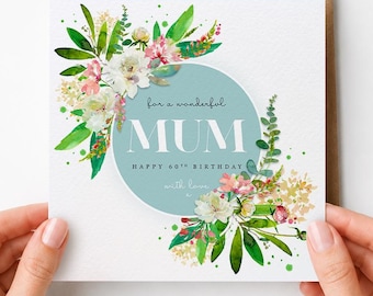 May include: A square greeting card with a light blue circle in the center featuring the white text "For a wonderful MUM HAPPY 60TH BIRTHDAY with love". The card is decorated with watercolor illustrations of green leaves and pink and white flowers.