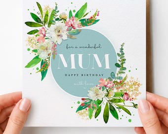 May include: A square greeting card with a white background featuring a central light blue circle. The text reads For a wonderful MUM HAPPY BIRTHDAY with love. The design is decorated with watercolor illustrations of green leaves and pink flowers.