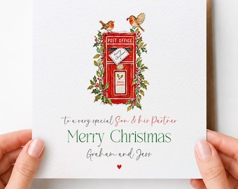 May include: A square white greeting card featuring a red post office box illustration surrounded by holly and two robins. Text reads: To a very special Son & his Partner, Merry Christmas, Graham and Jess, with a small red heart below.