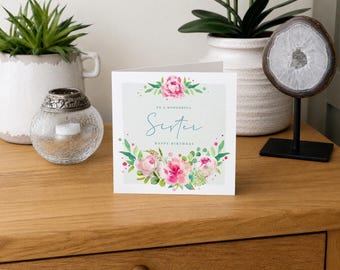May include: A square white greeting card featuring a watercolor floral design with pink peonies and green leaves. The text reads "TO A WONDERFUL Sister HAPPY BIRTHDAY" in blue and black lettering. The card is centered on a wooden surface.