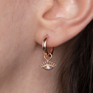May include: Rose gold hoop earrings with a dangling eye charm featuring a small clear rhinestone.