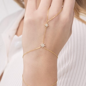 May include: A delicate gold hand chain bracelet featuring star-shaped accents with small, sparkling stones. The bracelet connects to a wrist chain, creating a unique and elegant jewelry piece. The design is visible against a white background.