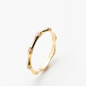 May include: A gold-colored ring with a bamboo design. The ring features small, clear gemstones set along the bamboo-like segments. The ring is set against a white background.