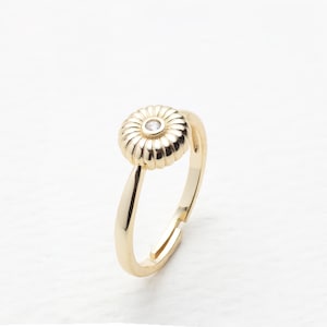 May include: A gold-colored ring with a flower-shaped design. The ring features a round, ribbed flower with a small, clear gemstone in the center. The band is adjustable and has a smooth, polished finish.