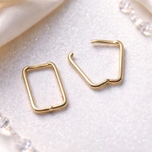 May include: A pair of gold-toned hoop earrings with a polished finish. One earring is shaped as a rectangle with rounded corners, while the other is shaped as a pentagon with rounded corners. Both earrings feature a thin, smooth metal wire construction.