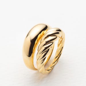 May include: Two gold rings. One ring is a smooth, rounded band. The other ring has a twisted rope design. Both rings are shiny and reflect light. The rings are set against a white background.