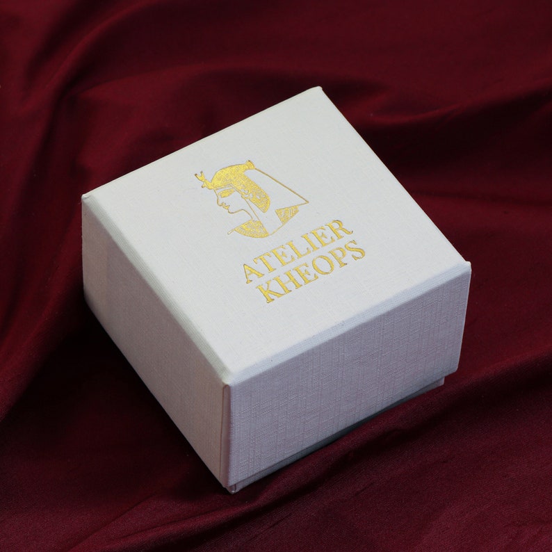 May include: A white square box with a gold logo and text that reads "ATELIER KHEOPS". The logo is a profile of a person with an Egyptian headdress. The box is sitting on a burgundy fabric.