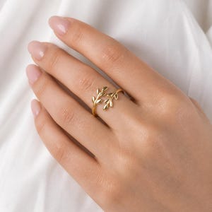 May include: A gold-colored ring featuring a delicate leaf design. The ring wraps around the finger, with two leaf clusters at the top. The ring is shown on a hand with a white background.