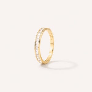 May include: A gold-toned band ring featuring a channel setting of small, clear baguette-cut stones along the outer surface. The interior of the band is clearly engraved with the text S925, indicating the metal composition of the jewelry piece.