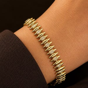 May include: A gold-colored bracelet with a spiky design, featuring small, sparkling stones set between the spikes. The bracelet is worn on a wrist, with a dark-colored sleeve in the background. The jewelry has a luxurious appearance.