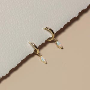 May include: A pair of gold hoop earrings with teardrop-shaped opal pendants. The earrings are displayed on a textured, off-white surface against a soft, neutral background. The opal pendants have a pearlescent sheen.
