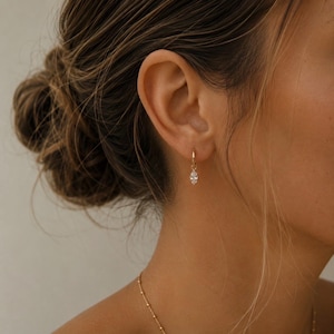 May include: A small gold hoop earring featuring a single marquise-cut clear gemstone charm dangling from the bottom. The earring is secured through the earlobe, showcasing a polished metallic finish and a faceted stone that reflects light.
