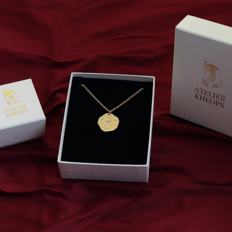 K&ouml;nnte beinhalten: Eine goldene Halskette mit einem gro&szlig;en, runden Anh&auml;nger, der ein detailliertes Design eines Auges zeigt. Die Halskette wird in einer wei&szlig;en Schachtel mit schwarzem Samtbezug pr&auml;sentiert. Die Schachtel tr&auml;gt den Schriftzug "Atelier Kheops" auf der Seite.