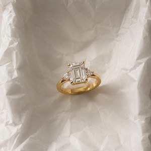 May include: A gold ring featuring a large emerald-cut center stone flanked by two smaller triangular-cut stones. The band is polished yellow gold, and the setting holds the three clear, faceted stones securely in a classic prong arrangement.