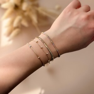 May include: A collection of gold bracelets adorns a wrist. The bracelets vary in design, including a delicate chain, a herringbone pattern, and a row of small, sparkling stones. The bracelets are displayed against a soft, neutral background.