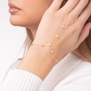 May include: A gold hand chain featuring a delicate design. The chain includes small, sparkling star and round accents. The jewelry is worn on the hand and connects to a bracelet. The chain is a stylish accessory.