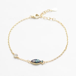 May include: Gold-toned bracelet featuring a small fish charm with iridescent blue and green mosaic-like inlay. A small, clear gemstone is set near the clasp. The bracelet is delicate and appears to be made of thin chain links.