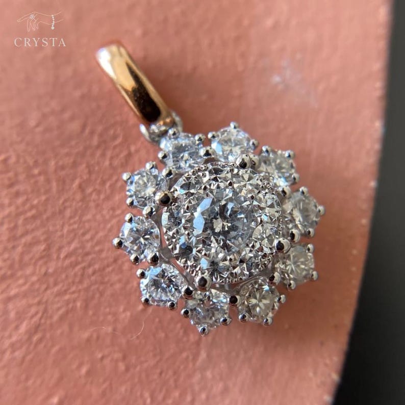 May include: A silver-toned pendant featuring a central round-cut clear gemstone surrounded by a halo of smaller round-cut clear gemstones in a floral arrangement. The pendant includes a polished gold-toned bail and the text CRYSTA.